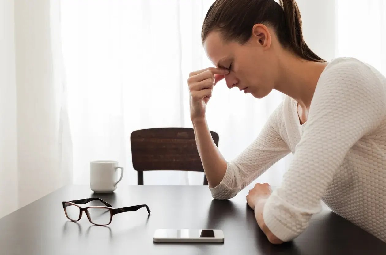Woman rubbing her tired eyes at a table, her glasses set down beside her phone